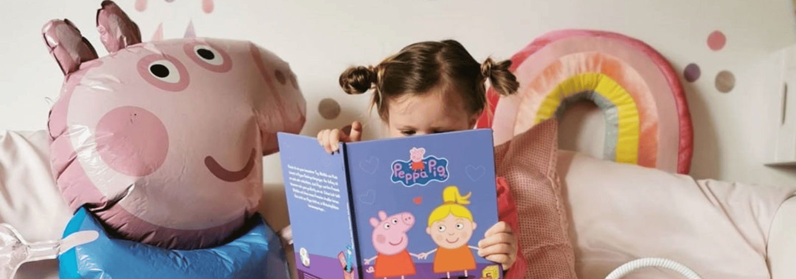 Ein kleines Kind mit Zöpfen sitzt auf einer Couch und liest ein Peppa Pig Buch, umgeben von einem großen Luftballon und einem Regenbogenkissen in einem gemütlichen, pastellfarbenen Zimmer.