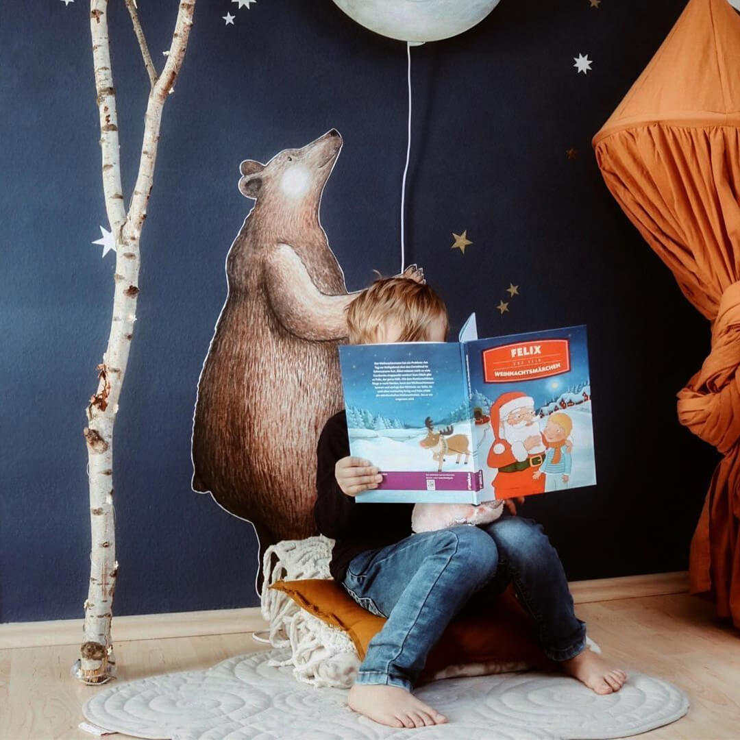 Ein Kind sitzt im Schneidersitz auf einem Kissen und liest in einem bunten Buch. Dahinter befindet sich eine Wandmalerei mit einem stehenden Bären und einer Birke, die mit Sternen und einem hängenden Mond geschmückt ist und eine fantasievolle und gemütliche Atmosphäre schafft.