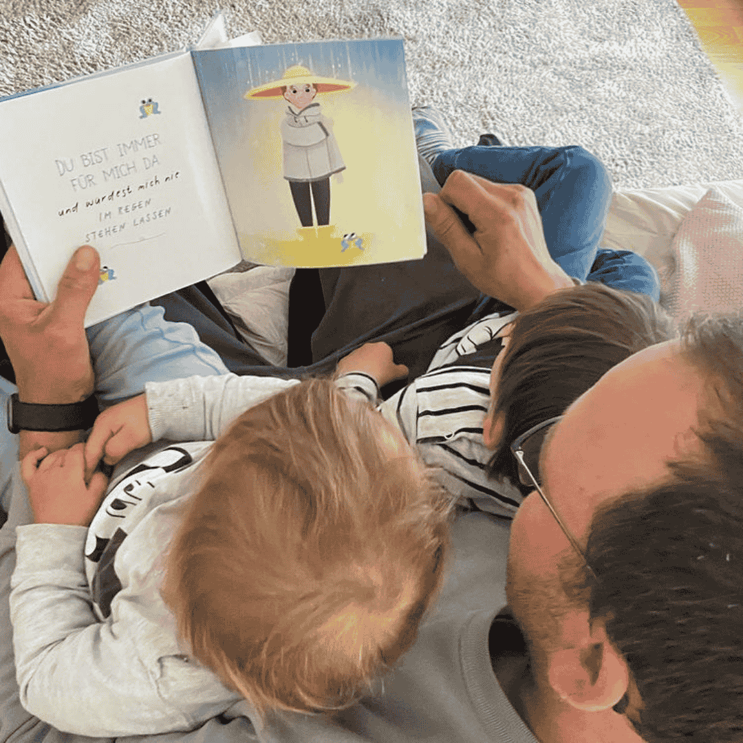 Ein Mann sitzt mit zwei kleinen Kindern auf seinem Schoß und liest gemeinsam ein deutsches Kinderbuch. Das aufgeschlagene Buch zeigt eine Illustration einer Person in einem gelben Kreis und einen Text auf der linken Seite, wodurch ein gemütlicher Familienmoment entsteht.