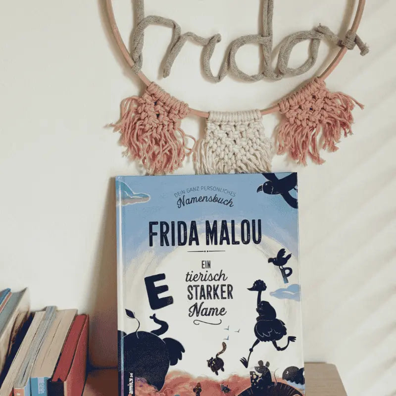 Ein Kinderbuch mit dem Titel FRIDA MALOU: Ein tierisch starker Name steht aufrecht in einem Regal zwischen Büchern. Dahinter verleiht ein dekorativer Reifen mit dem Namen Frida aus Garn und rosa Quasten der Szene einen besonderen Charme.