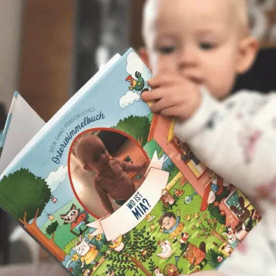 Ein Baby hält ein farbenfrohes Framily Wimmelbilder-Buch mit dem Titel "Dein Osterwimmelbuch mit Foto-Cover" in der Hand, das Tiere, eine Spielplatzszene und ein rundes Foto von dem Kind auf dem Umschlag zeigt.