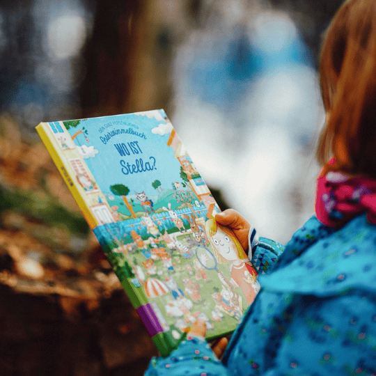 Ein Kind mit blauem Mantel und rosa Schal hält ein bunt illustriertes Bilderbuch mit dem Titel Wo ist Stella? in der Hand und schaut es an.