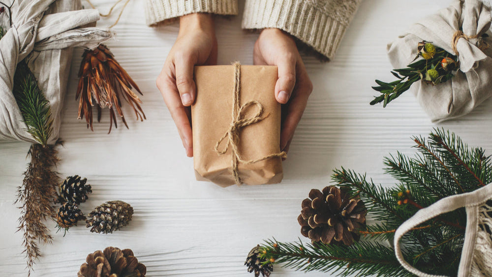 Person hält Geschenk mit braunem Geschenkpapier in der Hand, drumherum liegen Tannenzapfen und Tannenäste als Dekoration