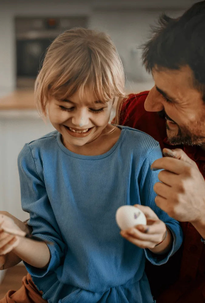 Ein lächelndes Mädchen in einem blauen Hemd hält ein Ei, auf das ein kleines Gesicht gemalt ist, während ein Mann neben ihr lacht und sie anschaut. Sie scheinen gemeinsam in einer Küche Ostereier zu verzieren.